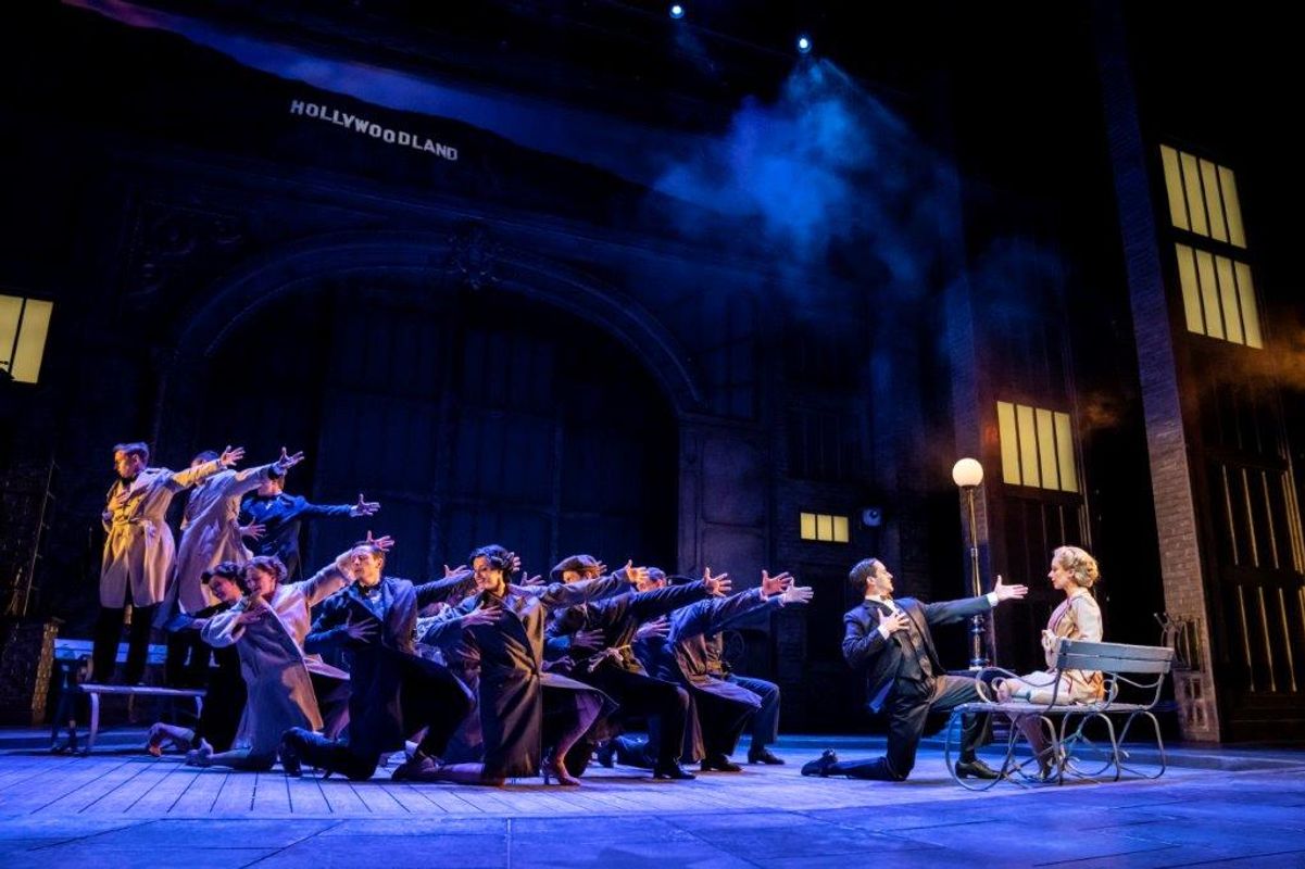 A woman sits on a bench with a man at her knees reaching his hand out to her, a group of dancers are doing the same behind the man