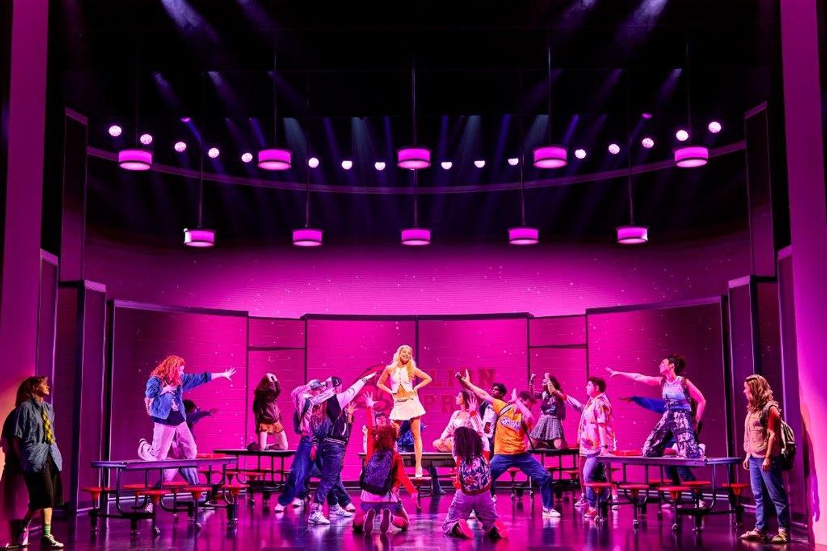 Stage performance with a Mean Girls cast member in a yellow dress standing on a table, surrounded by actors pointing at her. The vibrant pink lighting creates an energetic atmosphere.