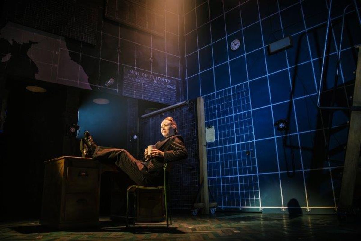 A person in a dimly lit room with a grid-patterned wall sits relaxed at a desk, feet up, holding a mug. The setting suggests an office-like environment.