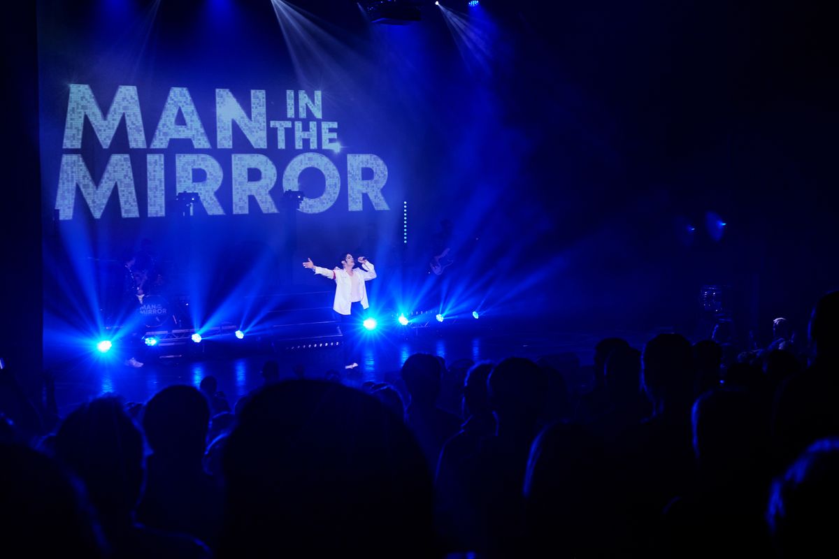 Performer on stage with "Man in the Mirror" projected behind. Blue spotlights illuminate the scene, creating a vibrant and energetic atmosphere.
