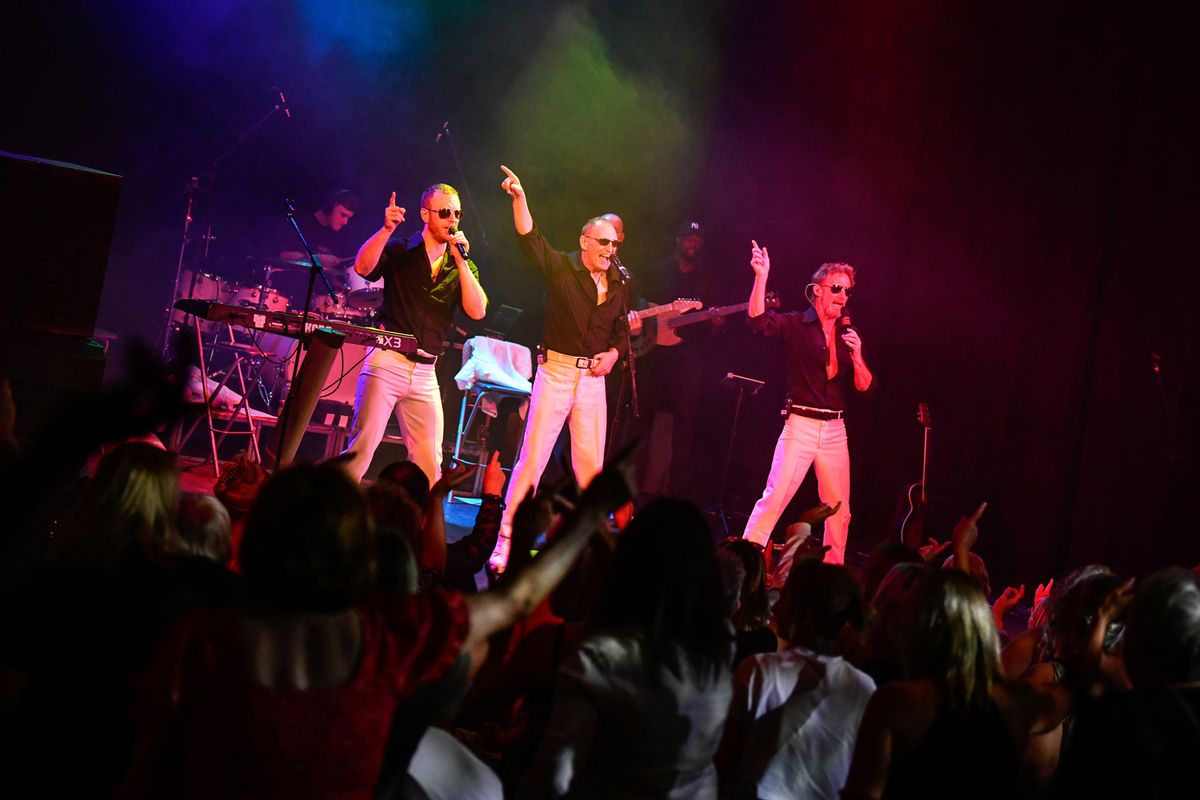 Three people dressed in white trousers, black shirts and sunglasses point their right arm in the air.