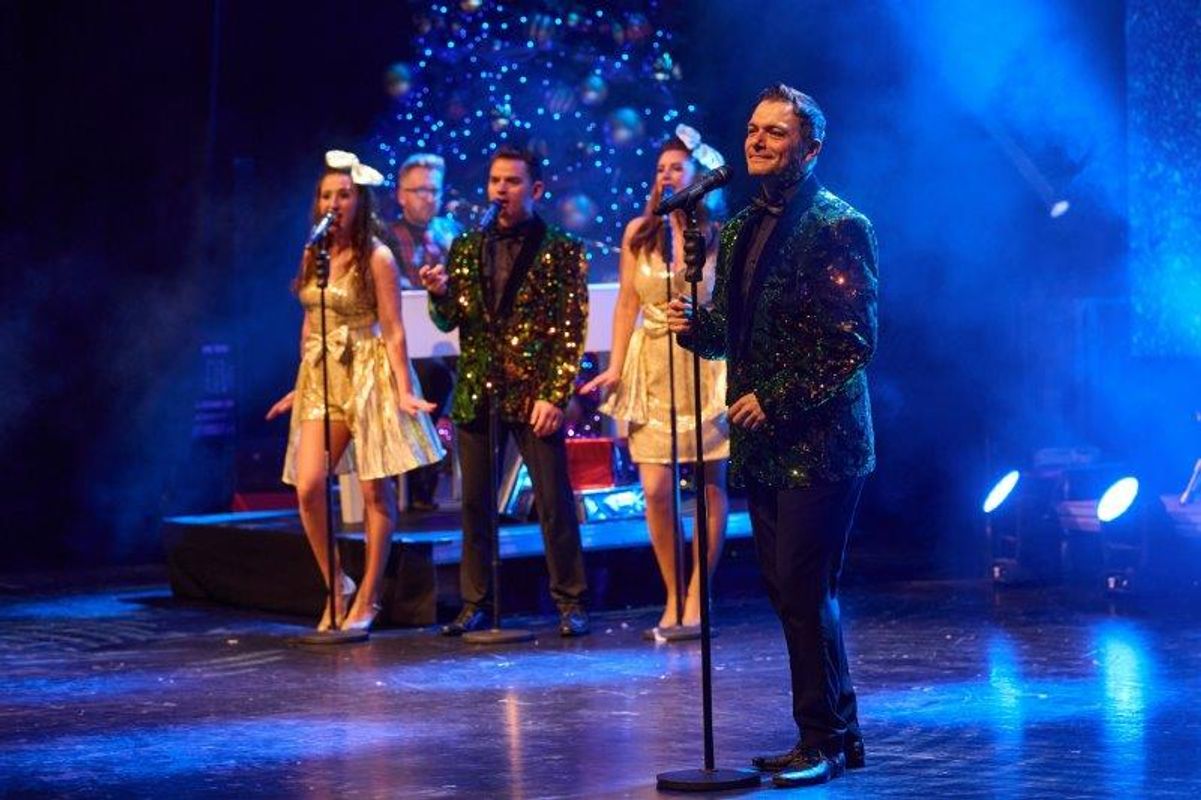 Four people, dressed in sparkly Christmas outfits, sing on stage.