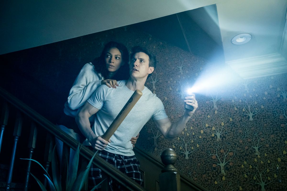 A man in pajamas holds a flashlight and a bat, looking anxious, with a woman clutching his arm. 