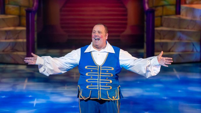 Buttons stands with his arms out to the side wearing a white collared shirt with a blue waistcoat.