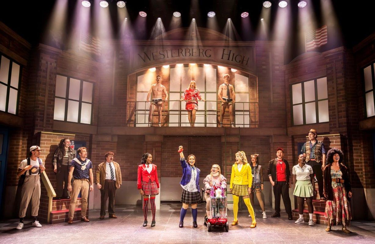 Stage performance at Westerberg High, featuring a diverse group of energetic actors in vibrant outfits, raising fists triumphantly, conveying excitement.