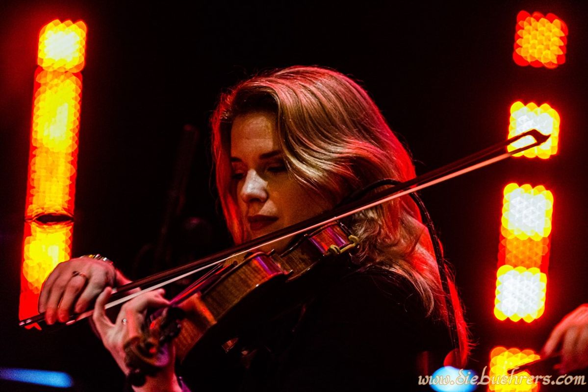 A woman with blonde hair plays a black violin on stage.