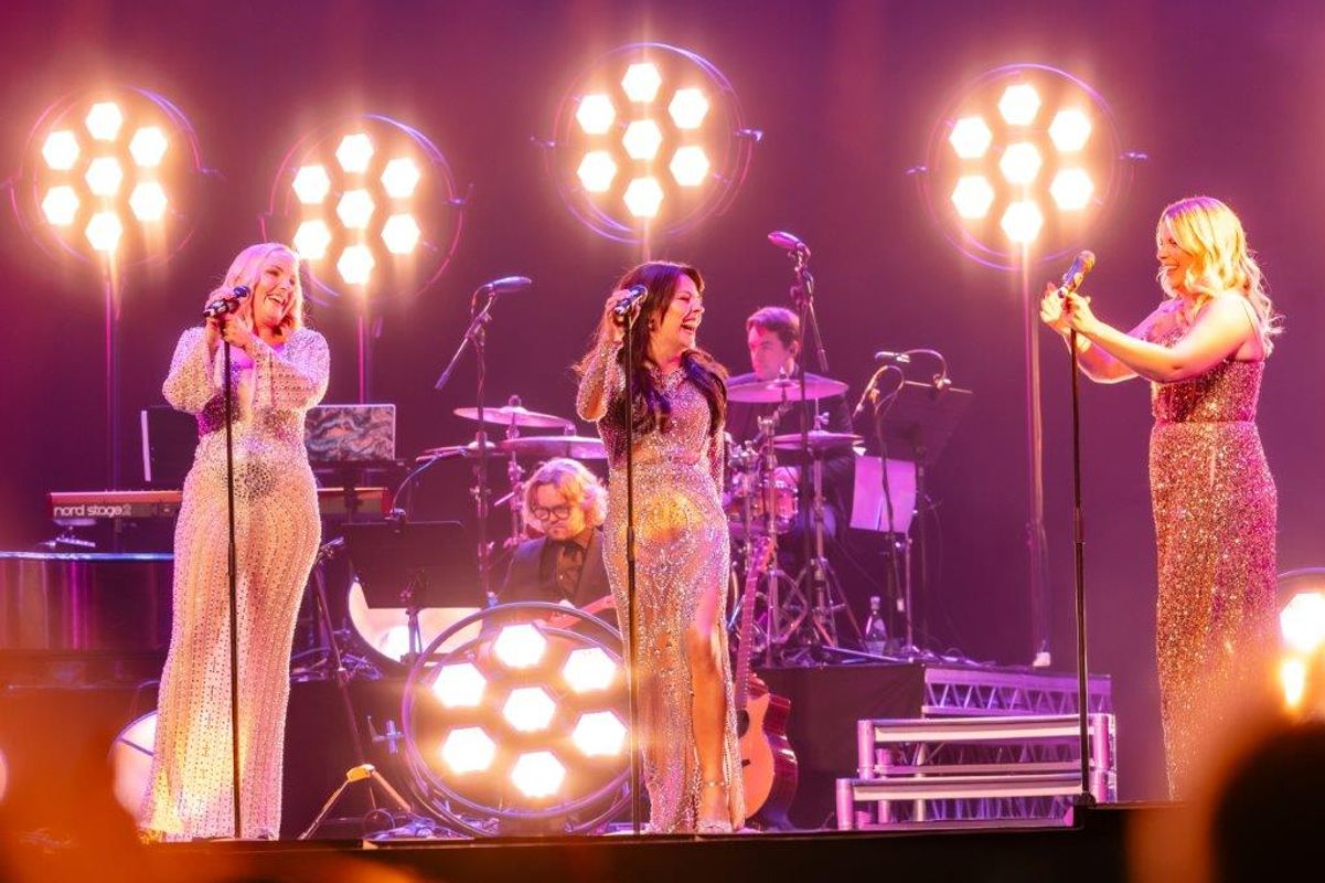 Kerry Ellis, Louise Dearman and Rachel Tucker in sparkling gowns sing joyfully on stage, surrounded by bright, circular lights. A band plays in the background, creating an energetic, lively atmosphere.