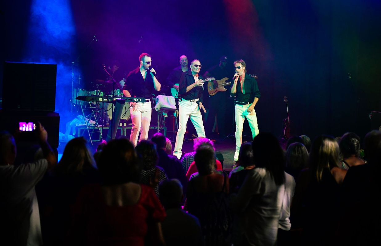 Three people dressed in white trousers, black shirts and sunglasses dance on stage.