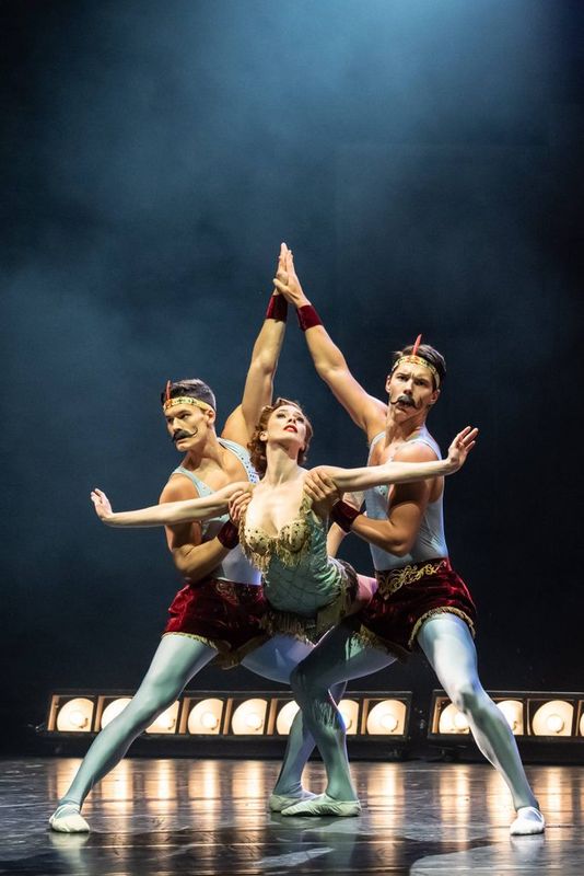 Two dancers raise their arms in a triangle motion with a third peering through.