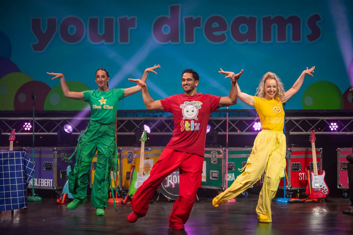 Three ensemble members, wearing colourful outfits, stand with their right leg kicking out.