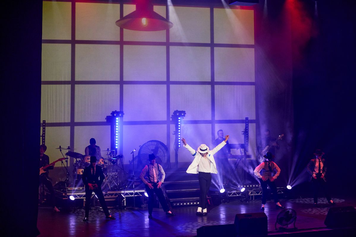 A dynamic stage performance with a dancer in a white suit and hat striking a pose center stage. Background features band members, colorful lighting, and dramatic shadows.