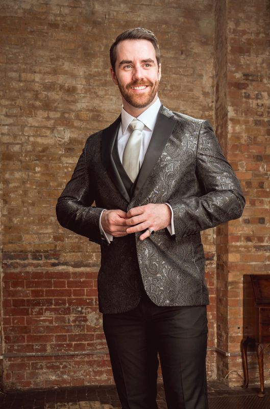 Luke McCall  in an ornate dark suit with a white tie stands against a textured brick wall, smiling confidently. The setting is rustic and elegant.