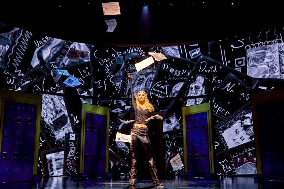 A person on stage throws papers into the air, surrounded by colorful lockers. The background displays chaotic black-and-white scribbles, creating a dynamic, energetic scene.
