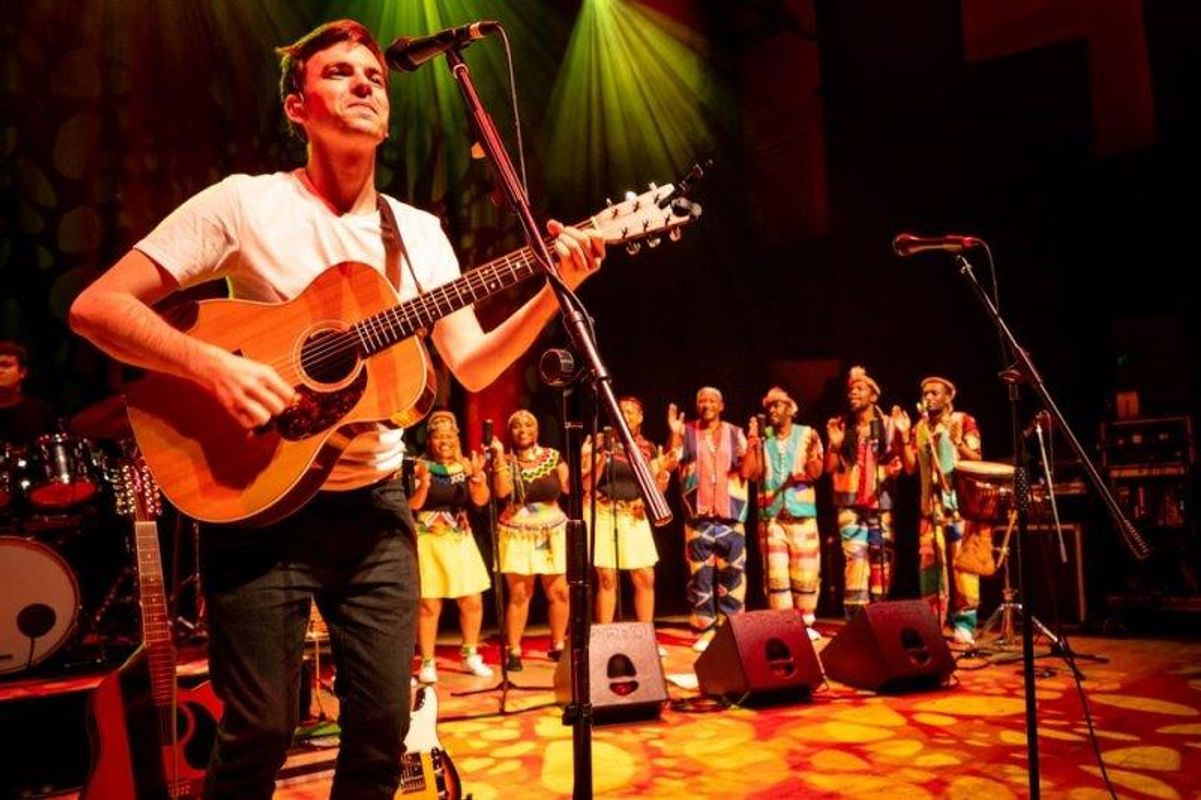The South African Cultural Choir are singing on stage with Paul Simon. They are all wearing bright and vibrant African clothing. Paul is playing a guitar