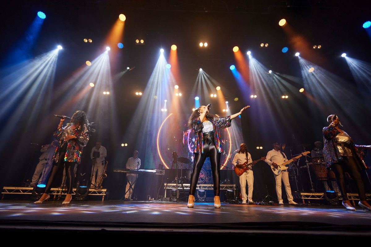 Three vocalists stand on stage wearing rainbow sequined blazers.