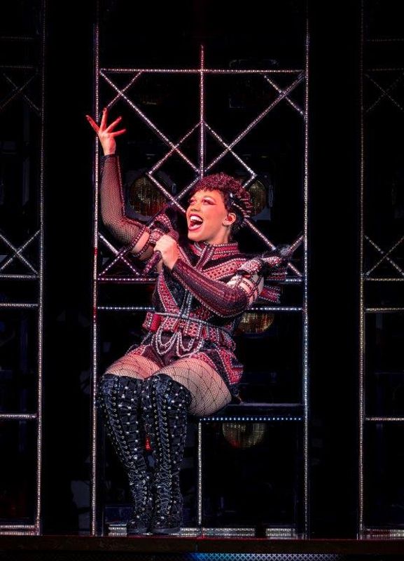 A person dressed in a pink and black studded outfit sits down with their right arm raised.