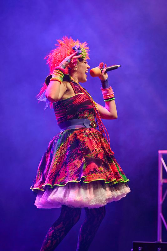 A person wearing a neon coloured dress and wig sings on stage.