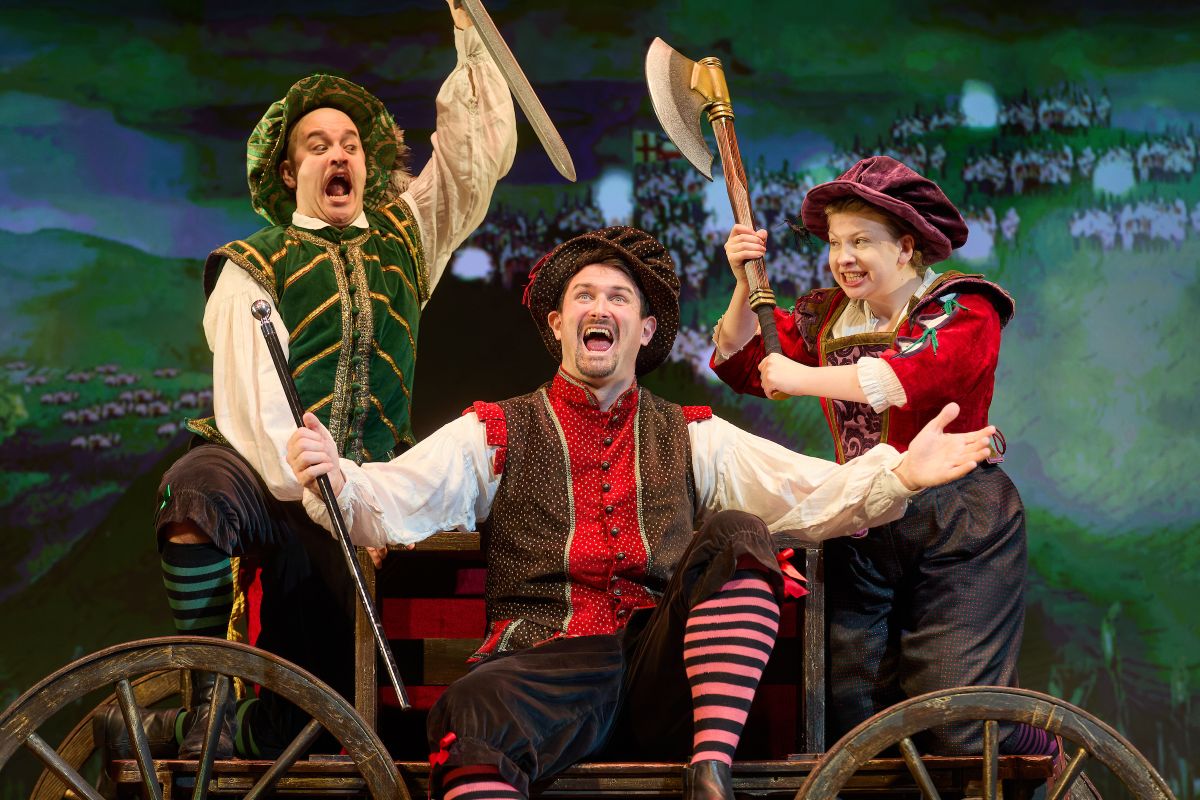 Three people on stage, they are all wearing 14th century clothing.  They are all stood on a cart with wheels holding weapons and a white flag