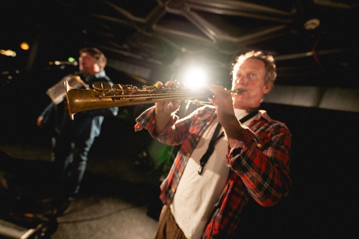 A musician plays a saxophone on stage, illuminated by bright lights. Another performer is visible in the background with a musical instrument.