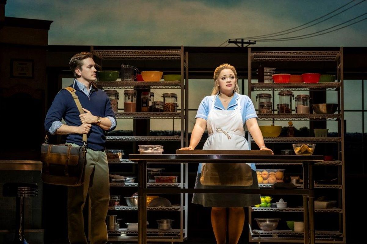 Two people are stood in a storage setting, one is behind the counter wearing a waitress uniform and the other is stood at the side holding a shoulder bag