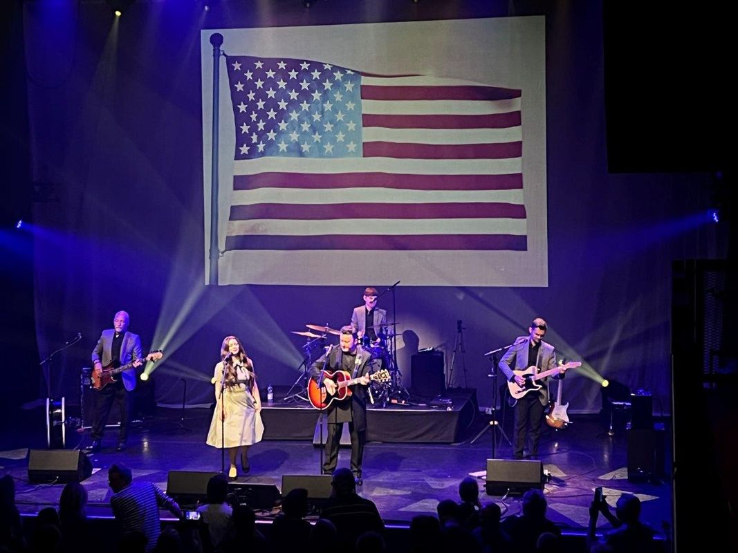 Johnny Cash Roadshow and band performing on stage. There is a large American flag behind them