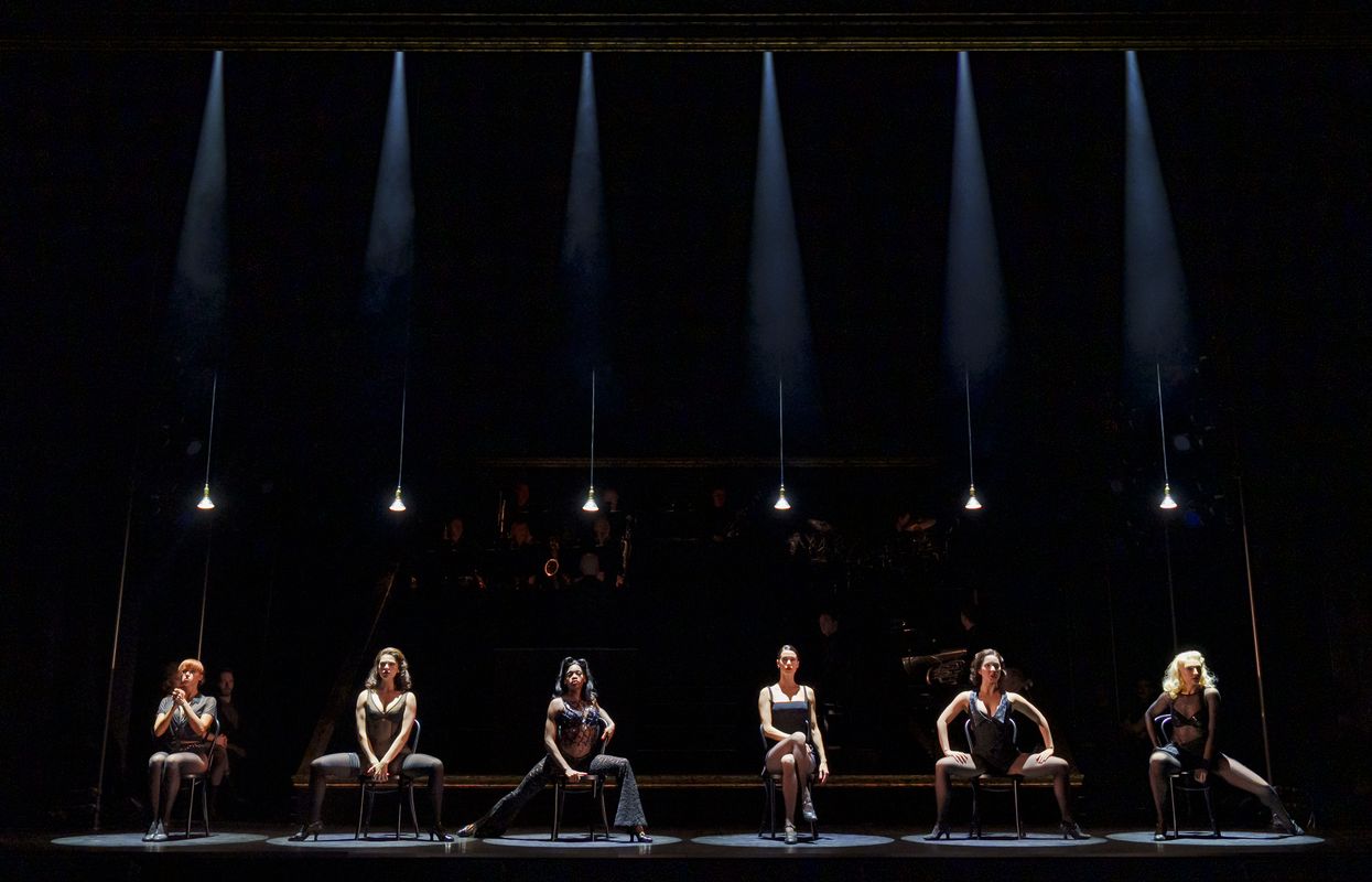 Six dancers on chairs in black skin tight mesh leotards and lingerie dancing on stage with low lighting