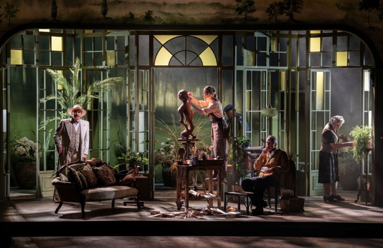 A theatrical stage scene depicting a lush, indoor setting with large windows. There are several individuals performing various activities: one adjusting a small statue, another sitting on a sofa, and others engaged in tasks. The design is rich with plants and period furniture, creating a vintage ambiance.