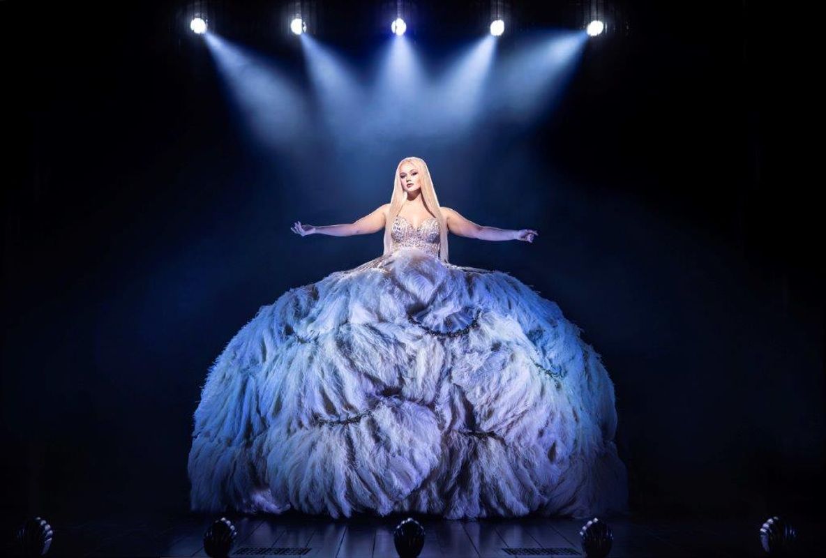 A lady with long blonde hair is wearing a glittery corset and a large feathered skirt