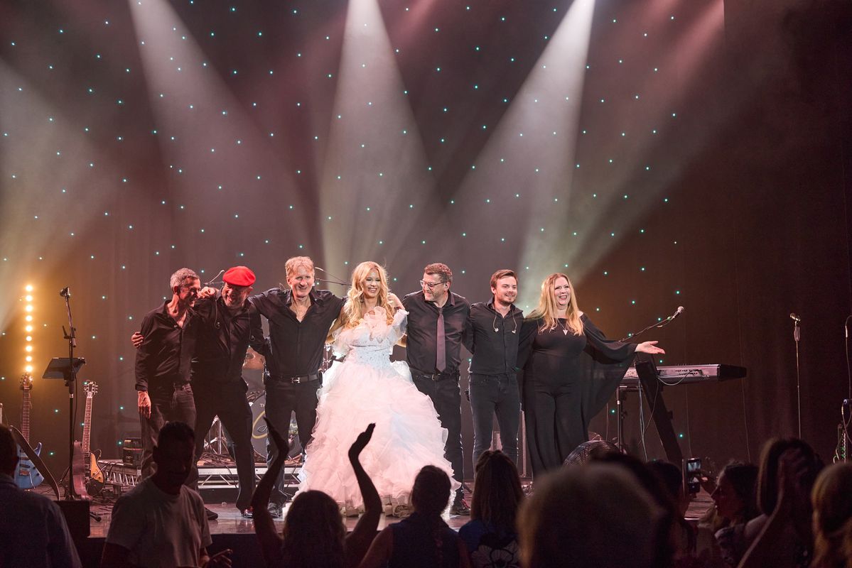 A group of performers stands on stage, holding hands and smiling at the audience. They are illuminated by stage lights, with musical equipment visible in the background. The audience is applauding.