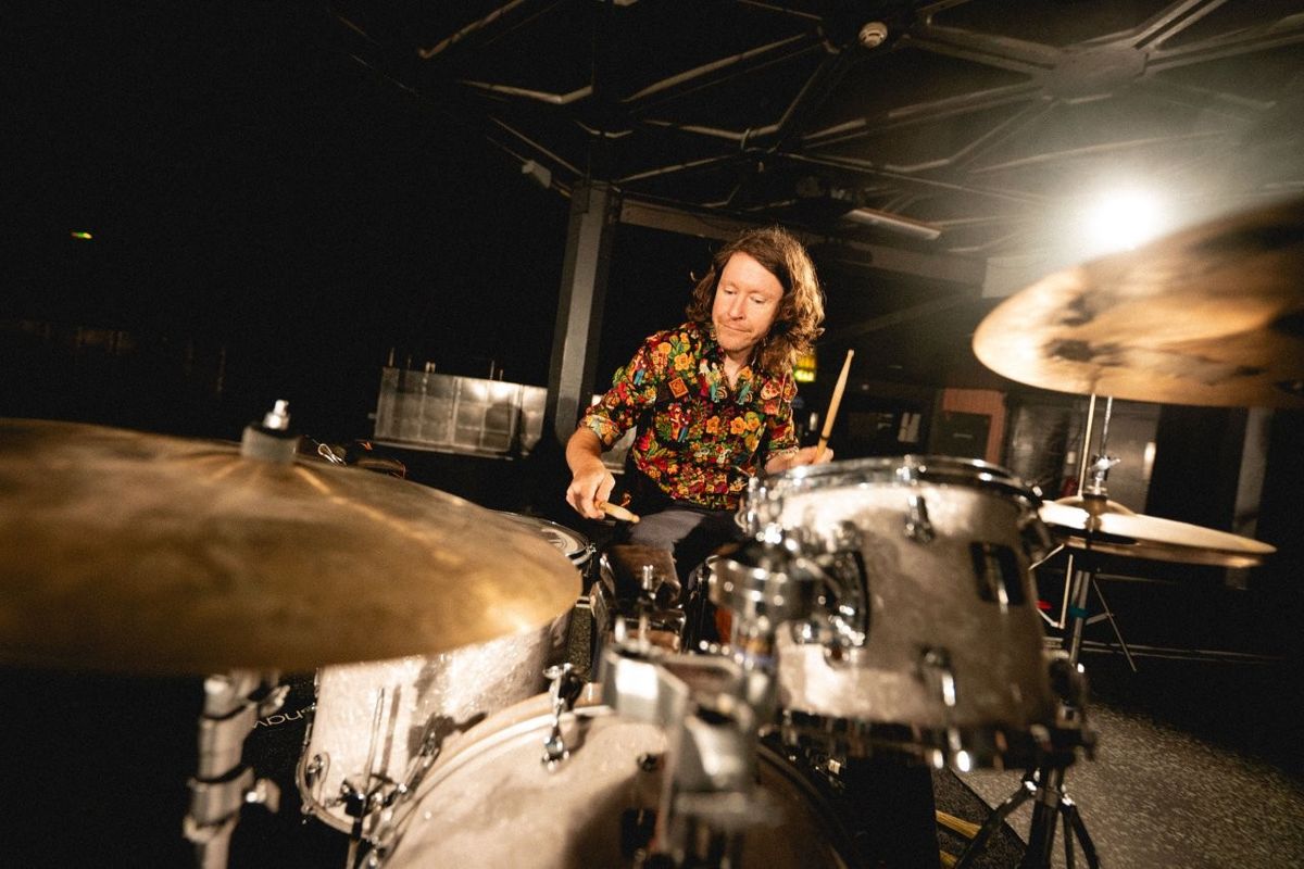 A person with long hair is playing a drum set in a dimly lit space. They are wearing a colorful floral shirt. The light softly illuminates the scene, creating a dramatic atmosphere.
