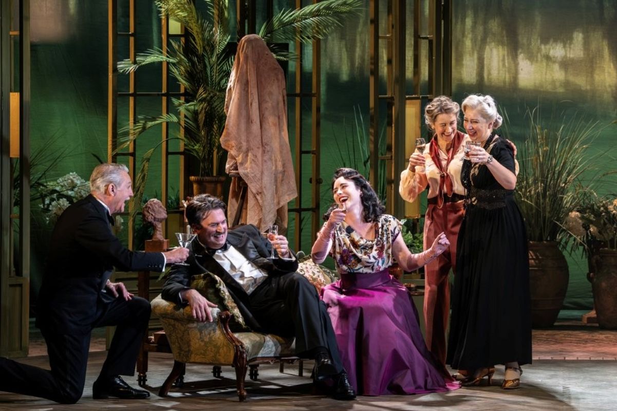 A group of people dressed in formal attire are cheerfully raising their glasses in a toast on stage. The set features lush greenery and elegant decor, creating a sophisticated ambiance.