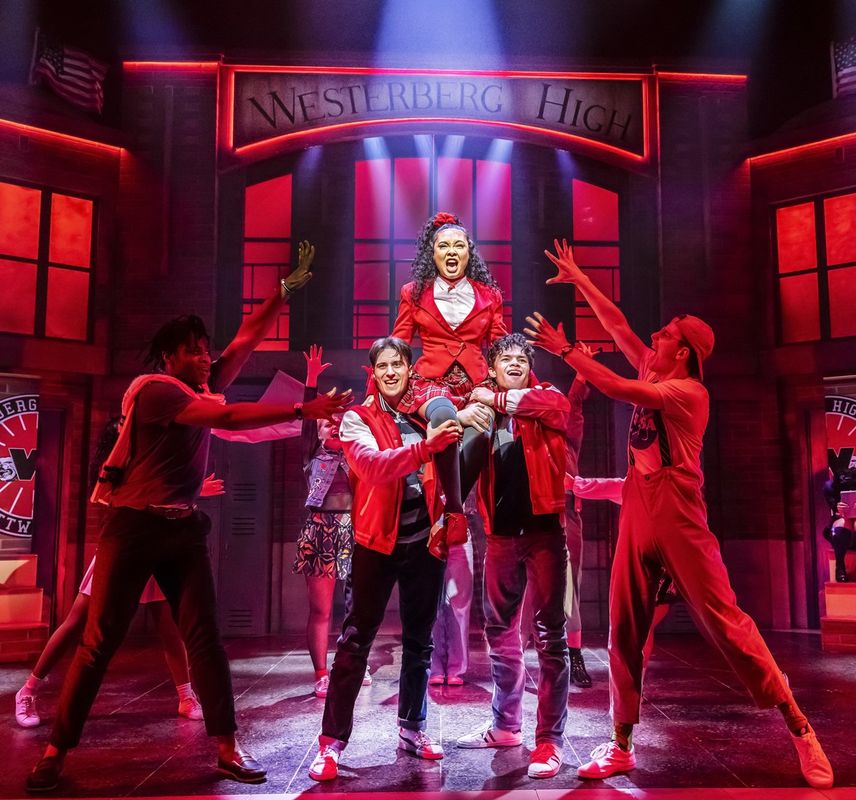 Stage performance of "Heathers: The Musical" at Westerberg High. Central figure in red blazer is energetically lifted by students in red jackets. Mood is vibrant.