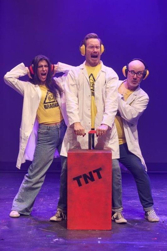 Three people wearing ear defenders press a giant TNT plunger.