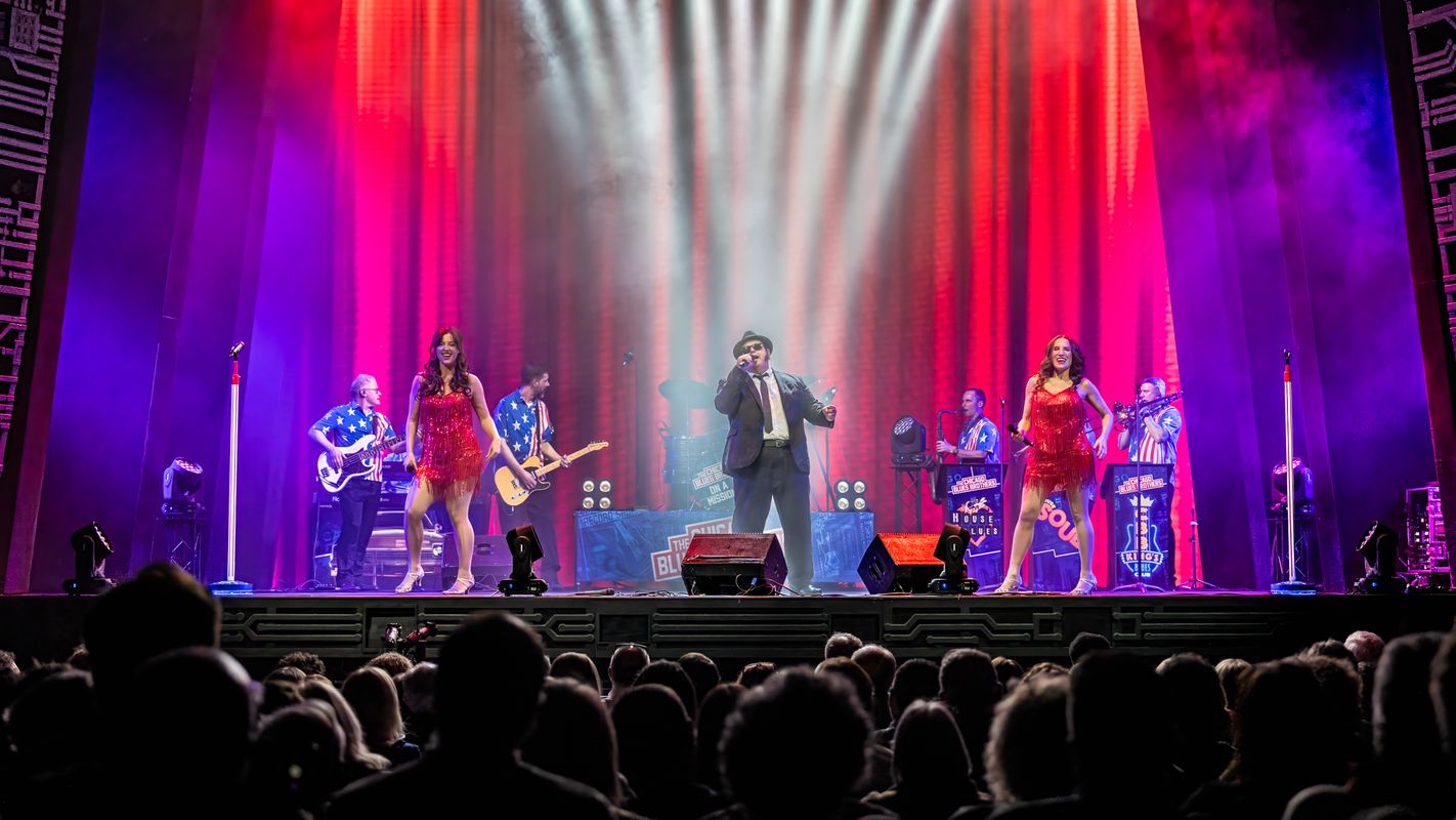 A person stands centre stage in a black suit and bowler hat accompanied by two dancers.