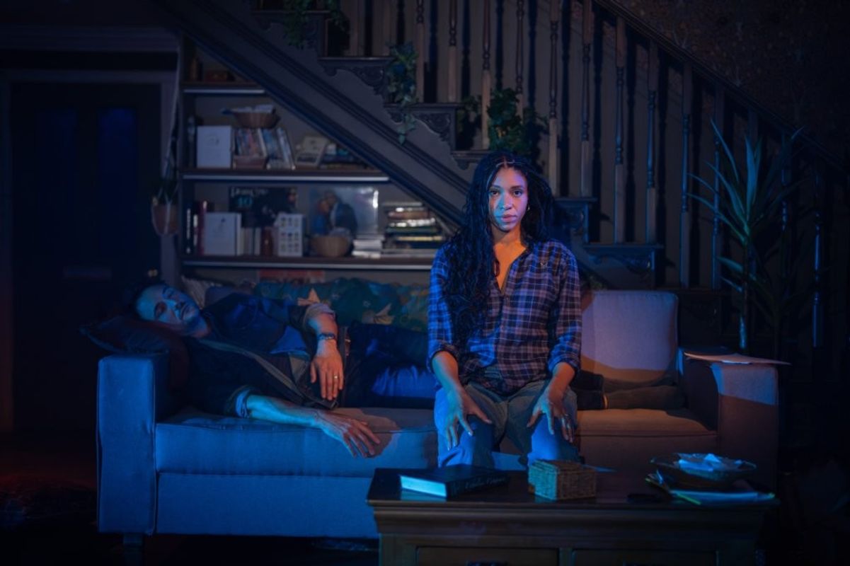 A woman is sat staring a head. A man is sleeping on the sofa next to her.