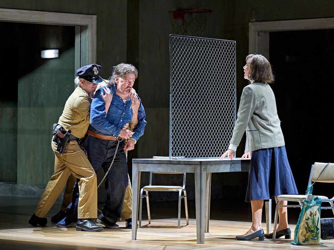 An inmate wearing handcuffs is being pulled away by prison guards. A lady is stood on the other side of the table watching