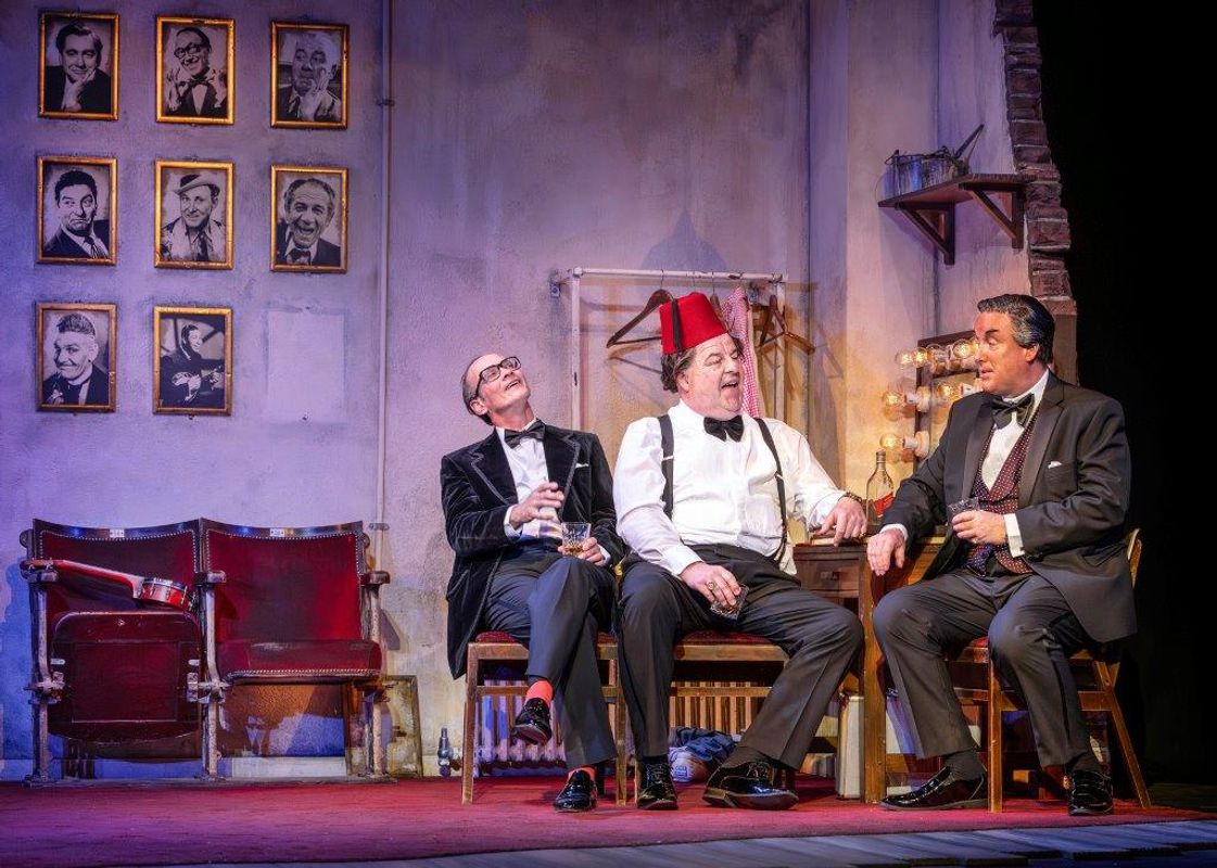 Three men sat together talking in a dressing room backstage type setting, one is wearing a fez and they are all dressed smartly
