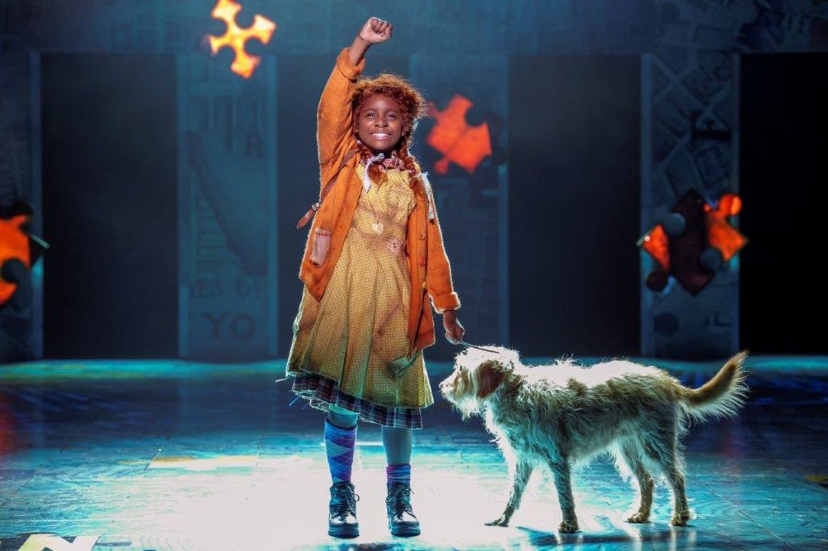 A young girl on stage, wearing an orange coat and yellow dress, raises her arm confidently beside a scruffy dog. The background features puzzle piece shapes.