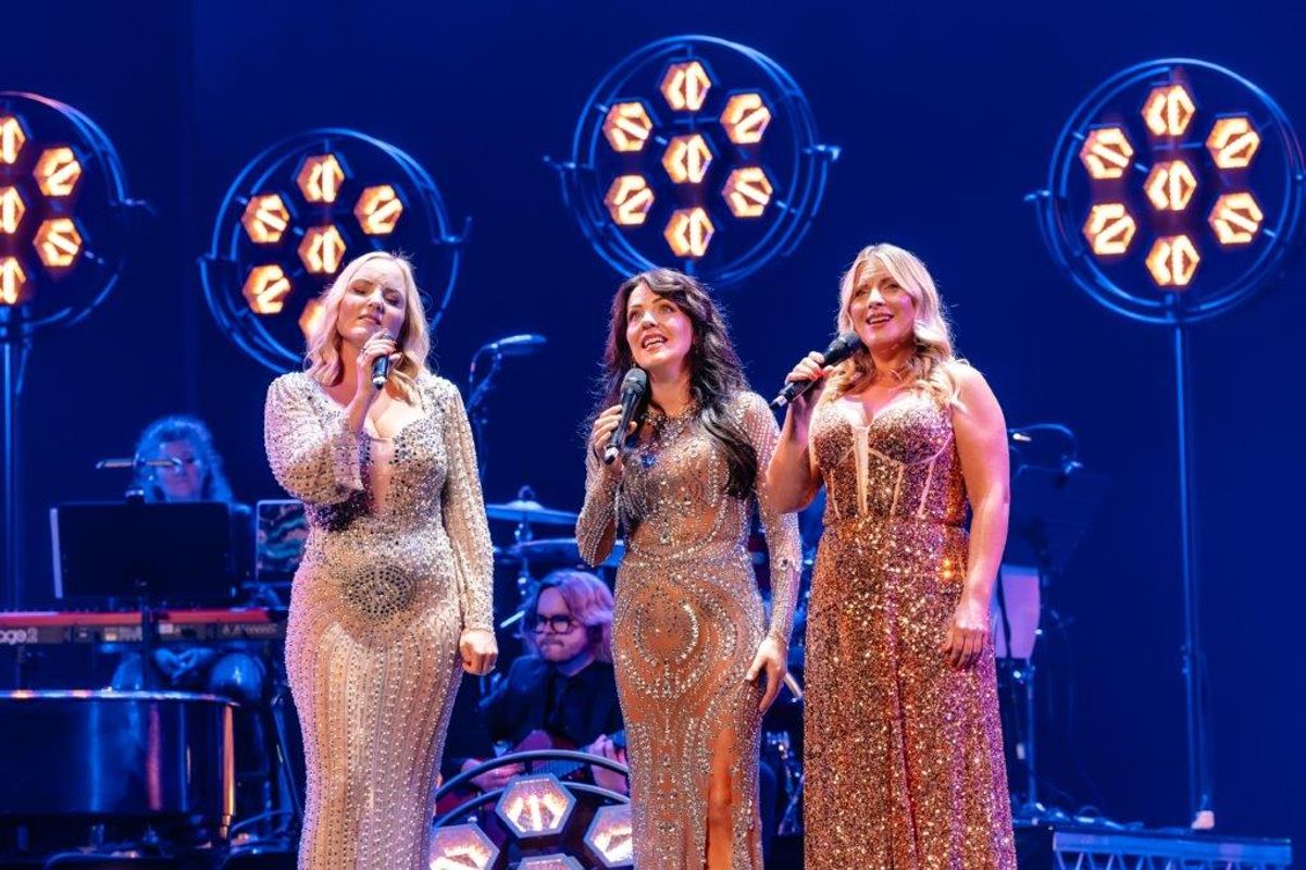 Three women in sparkling dresses sing on stage under warm, circular lights. A pianist and other musicians are faintly visible in the dimly lit background.