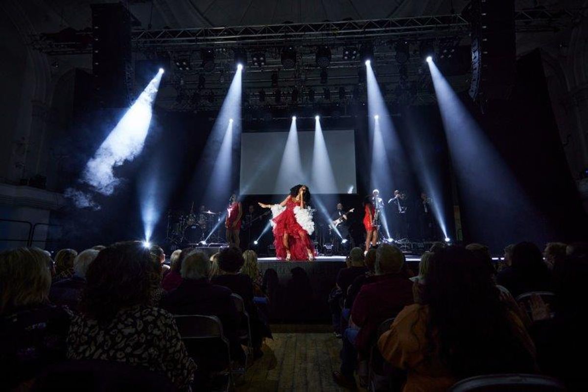 A person is stood centre stage wearing a red dress and white shawl.