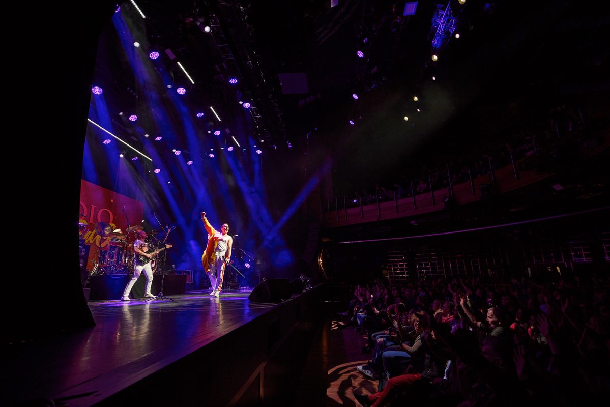 Performers singing and playing instruments on stage to an audience, bright lights are shining on the band