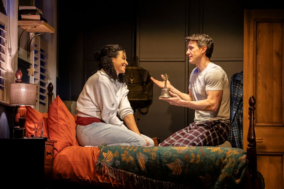 A woman and a man are sitting on a bed in a cozy, warmly lit room. The man, in pajamas, holds a small silver bell, engaging the woman in conversation. She wears a comfortable sweater and smiles at him, creating a sense of intimacy and warmth. The room has a patterned blanket and a softly lit nightstand.
