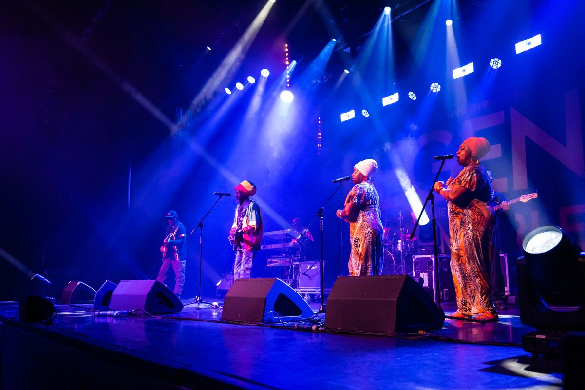A vibrant stage performance shows three performers in colorful attire and white hats singing into microphones. Blue and purple lights create an energetic atmosphere.