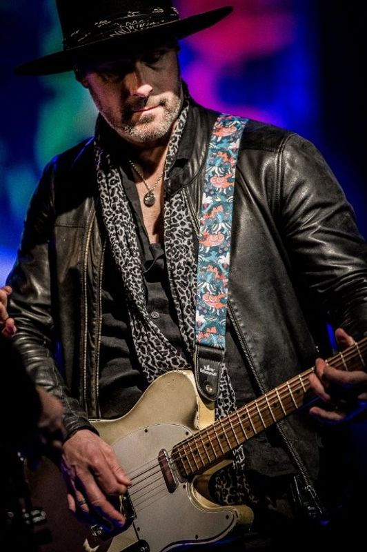 A musician in a leather jacket and hat plays a guitar with a colorful strap on stage against a backdrop of vibrant lights.