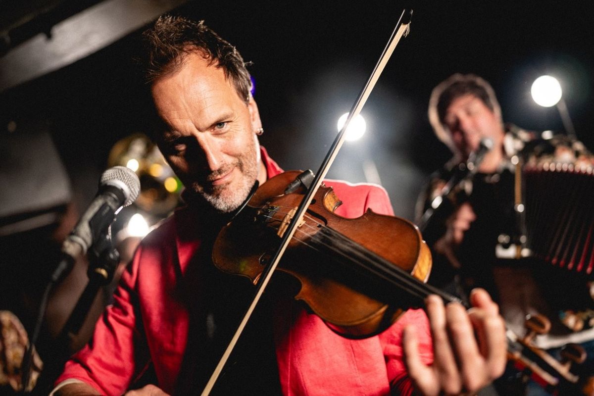 A man performing on violin during a live music session, wearing a bright red shirt under stage lighting.