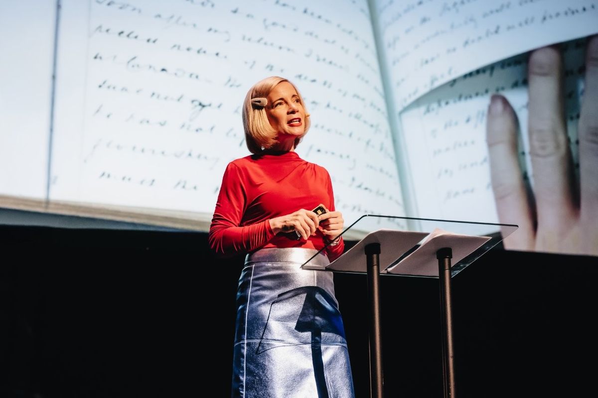 Lucy Worsley talking on stage. She is wearing a red top and a silver pencil skirt.