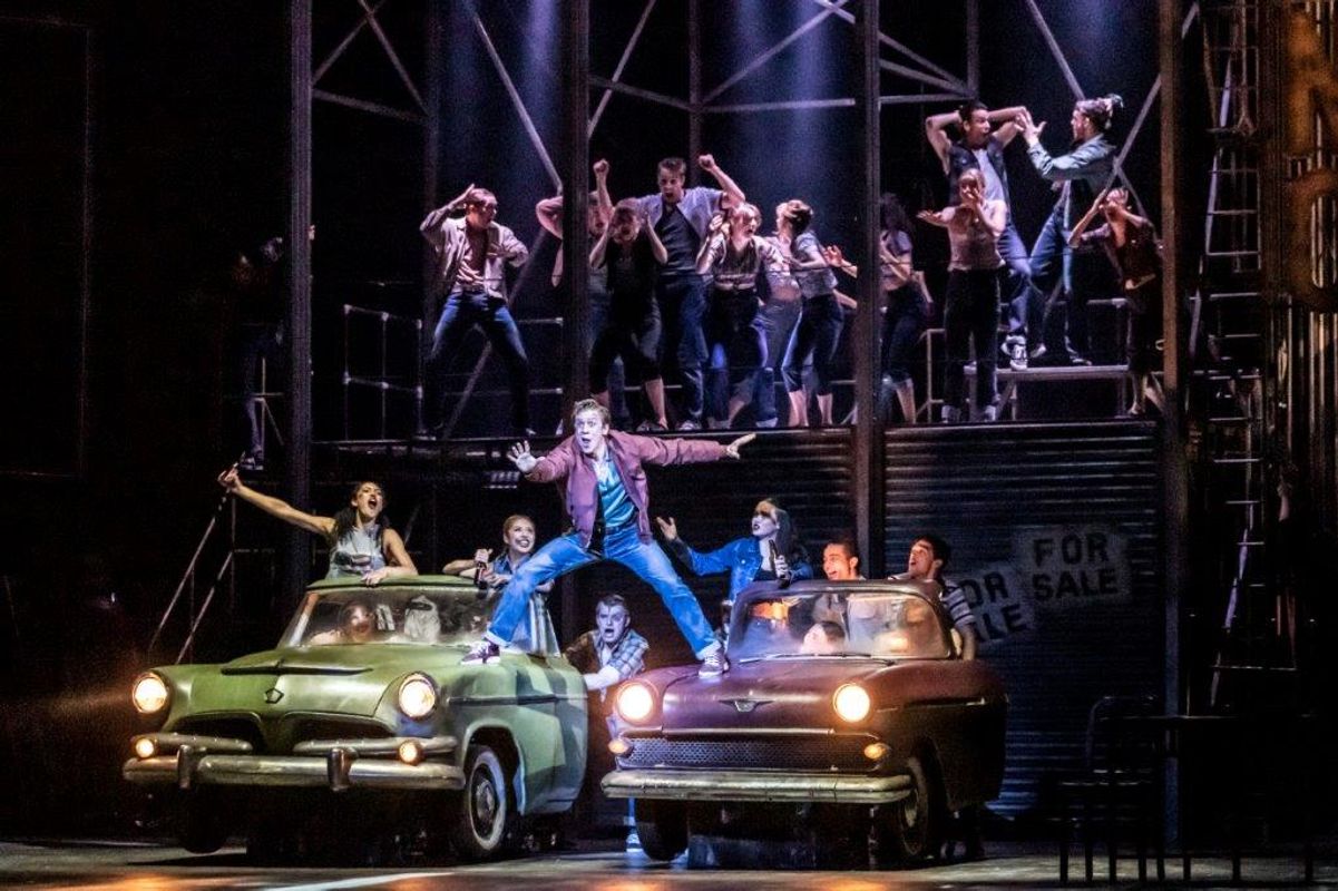 A dynamic stage scene with a performer in a red jacket and blue jeans energetically dancing on cars. A group of dancers energetically poses around, with dramatic lighting and an industrial backdrop creating an intense, lively atmosphere.