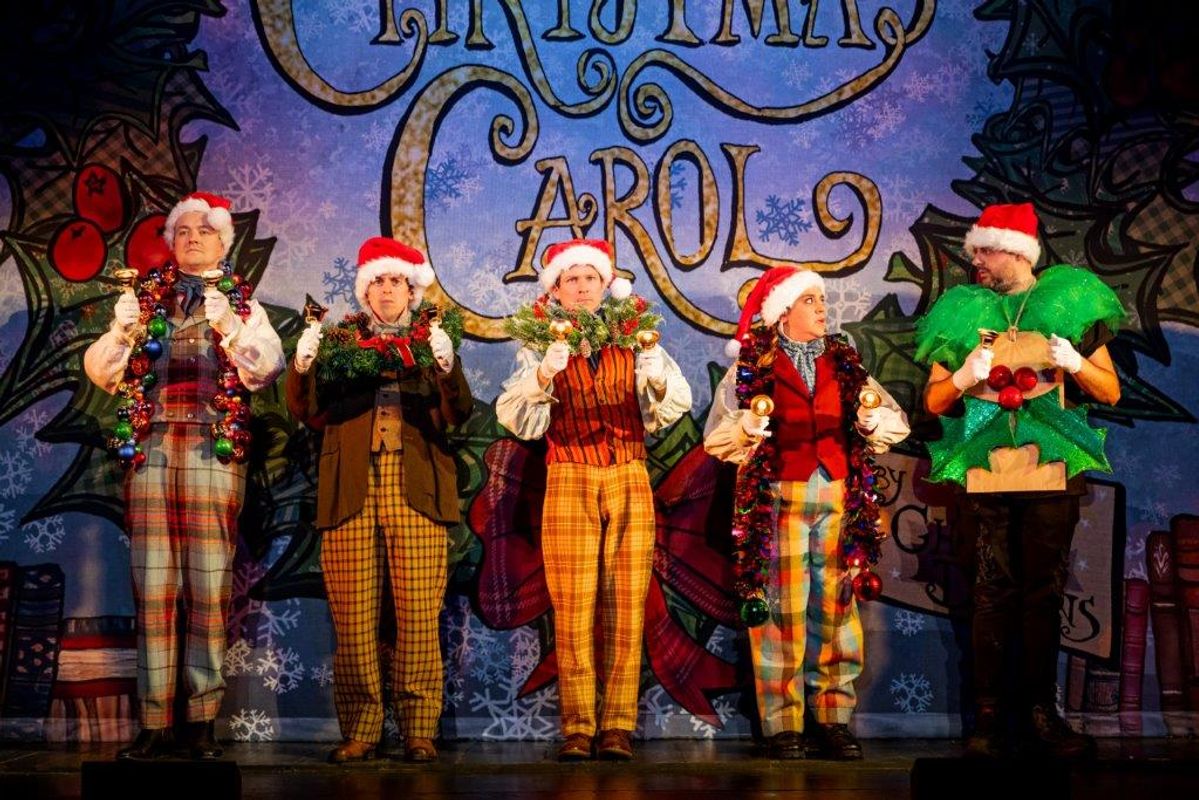 Five people dressed in festive outfits play bells on stage.