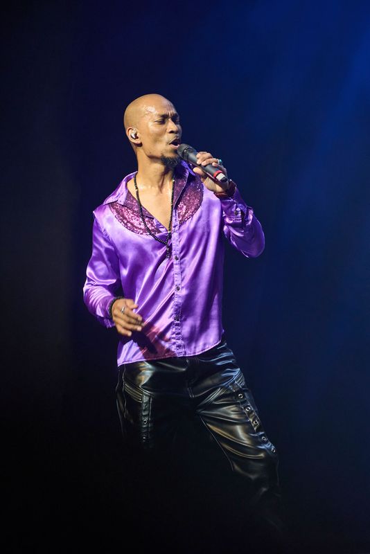 A person wearing a purple shirt with sequined collar and black trousers, holds a microphone.