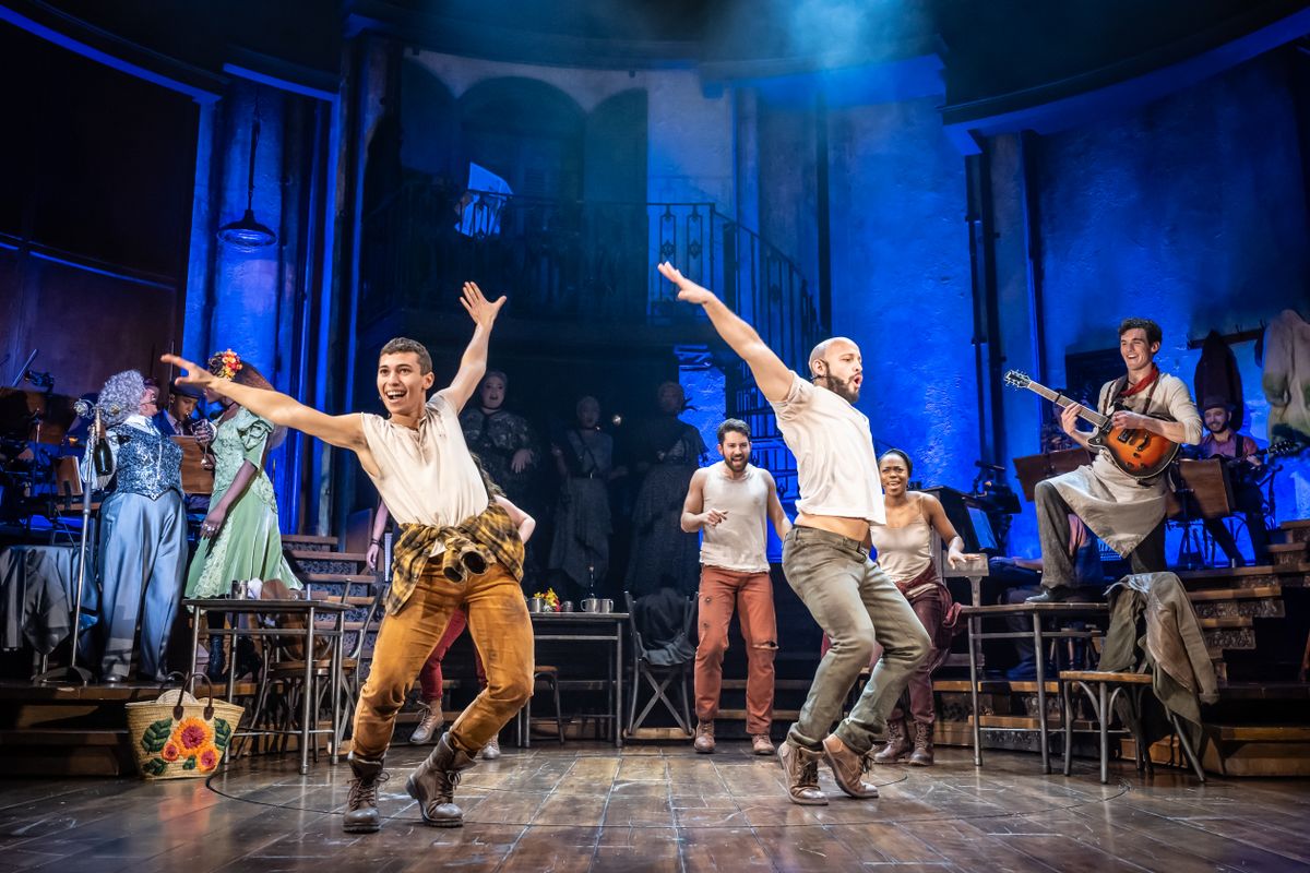 Two dancers stand centre stage with there knees bent and arms in a flying position surrounded by ensemble and muscians.